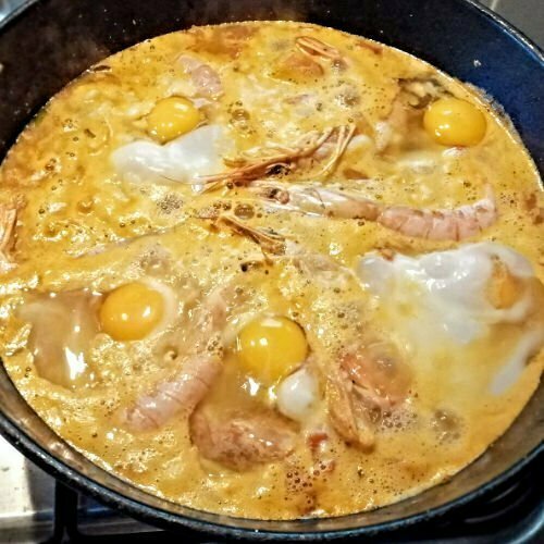 A huge pot of arros caldoso con mariscos simmers on a kitchen stove. Egg and large prawns are sitting on top.