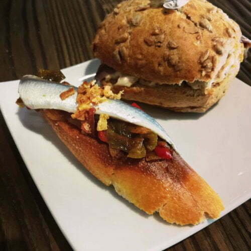 Boquerones fillet on a small slice of bread, served on a plate with a larger bread roll in the background