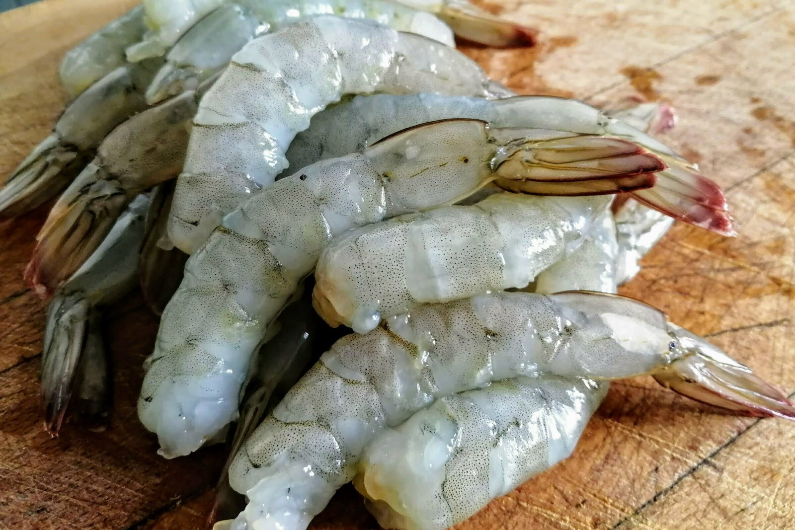 A stack of large uncooked prawns sit on top of a wooden chopping board. 