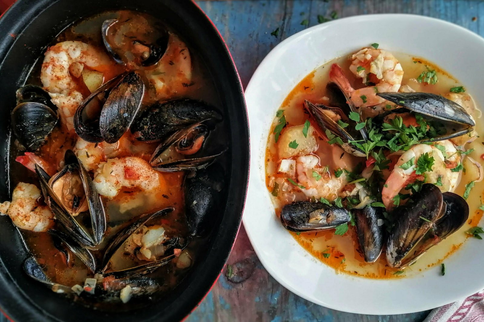 A large red pot of Spanish seafood. stew sits beside a white bowl, filled with prawns, fish, and museels in a red paprika infused broth. 