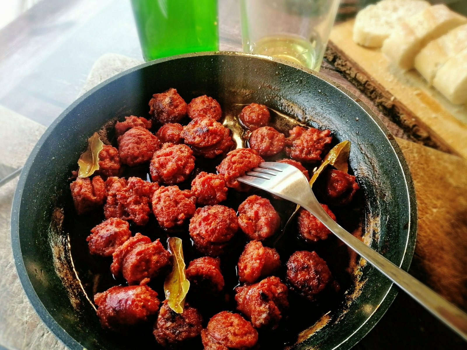 A pan of chorizo cooked in cider sits on a rustic chopping board with some apple cider oured in a glass sitting nearby