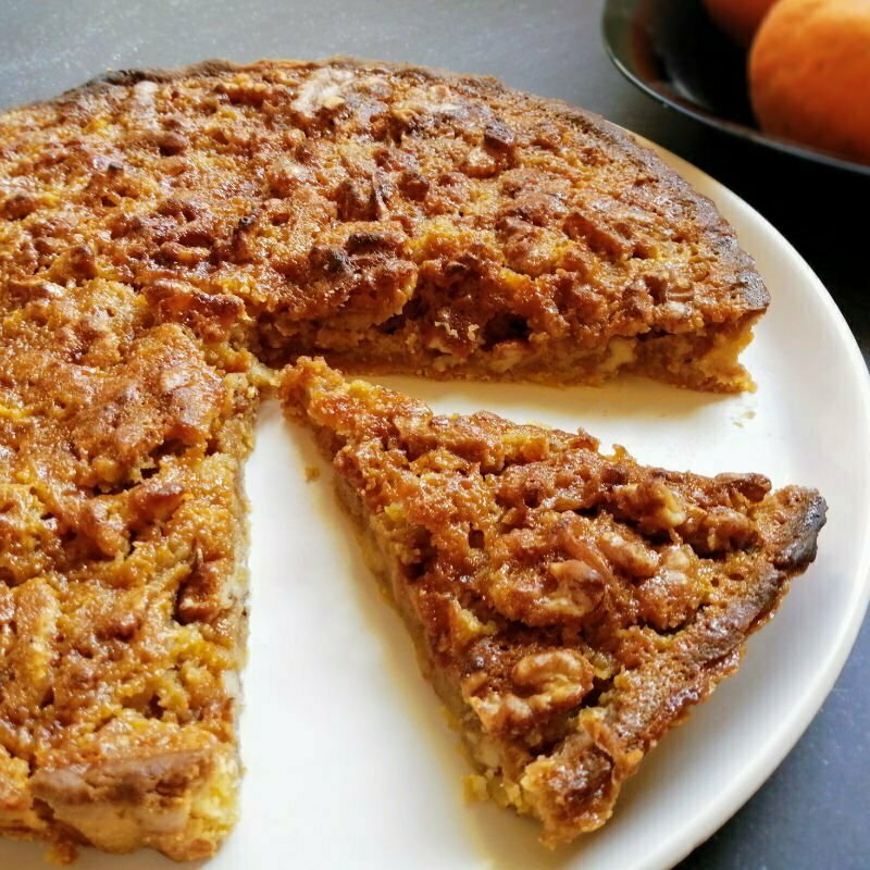 A slice of walnut tart sits on a white plate on a granite countertop