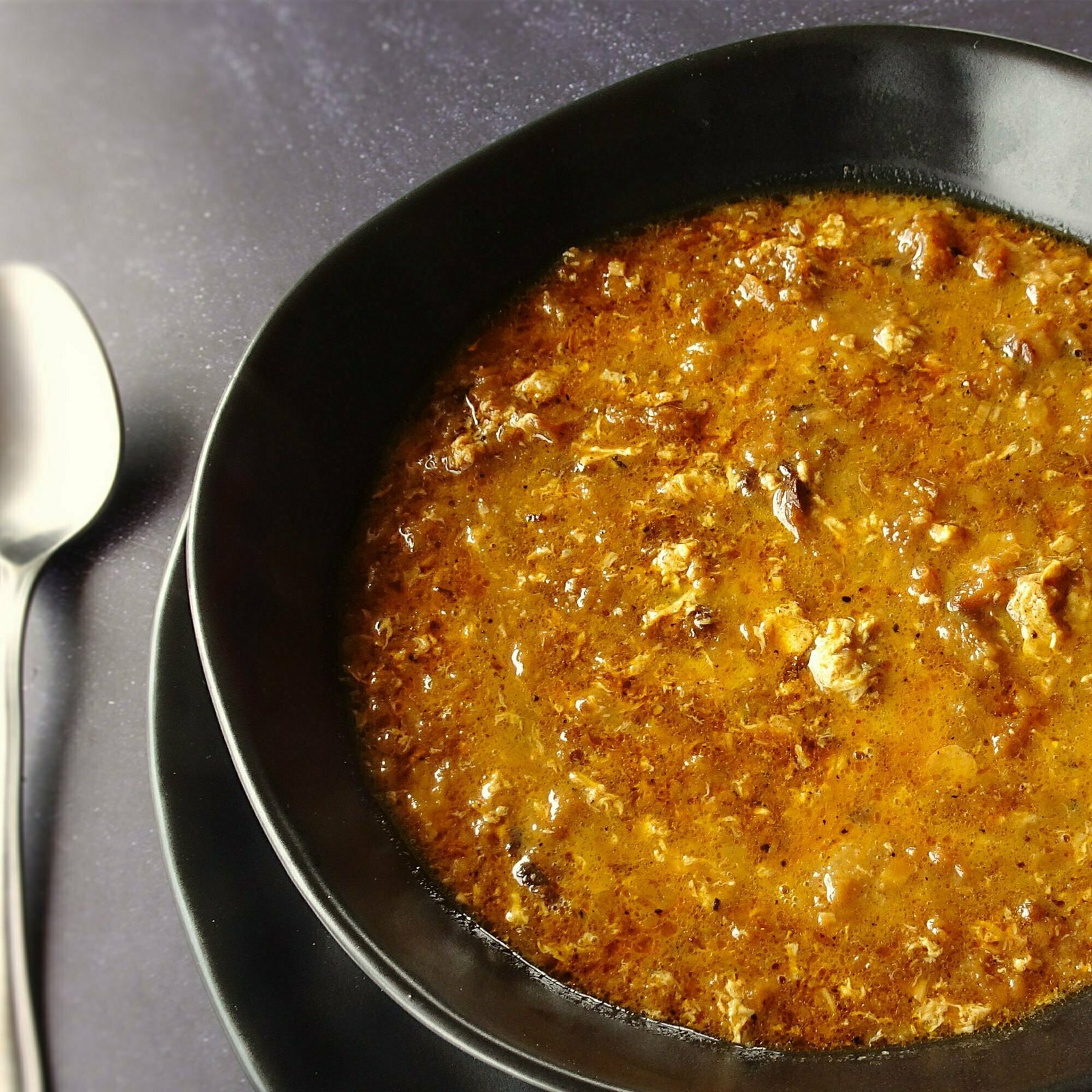 A bowl of garlic soup sits on a tabletop beside some slices of toasted bread and a silver spoon