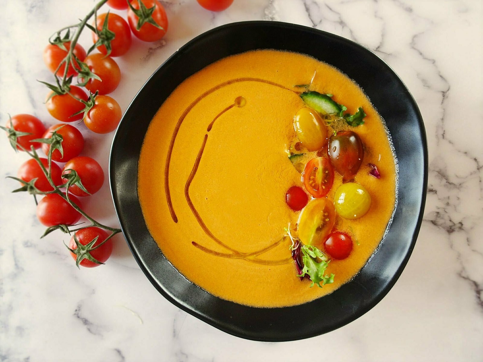 a bowl of gazpacho andaluz sits garnished with fresh salad andon a marble coutertop beside some ripe cherry toatoes