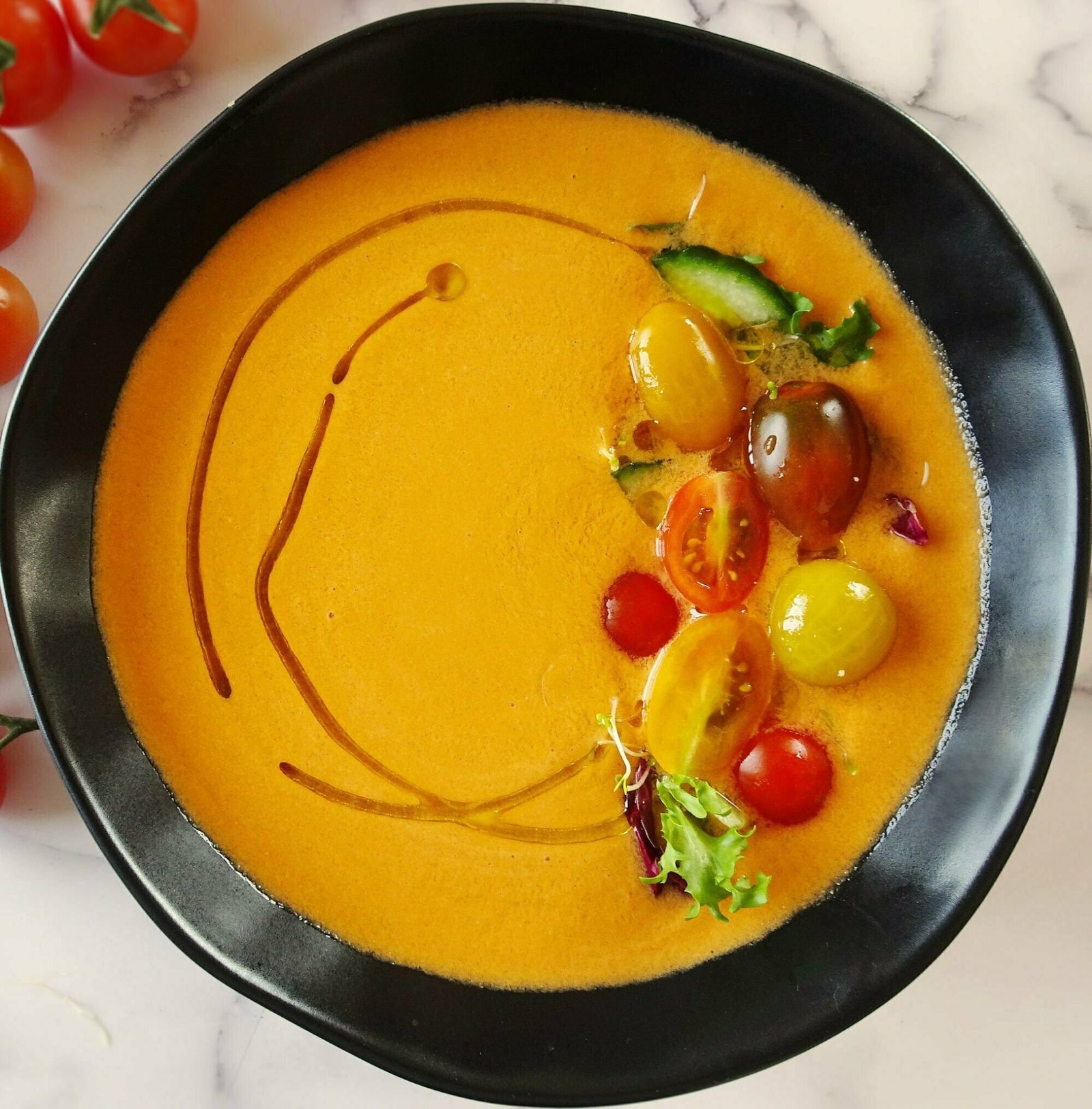 a bowl of gazpacho andaluz sits garnished with fresh salad andon a marble coutertop beside some ripe cherry toatoes