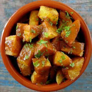 an earthenware dish sits on a blue wooden table and is filled with golden friend potato pieces smothered with Spanish bravas sauce