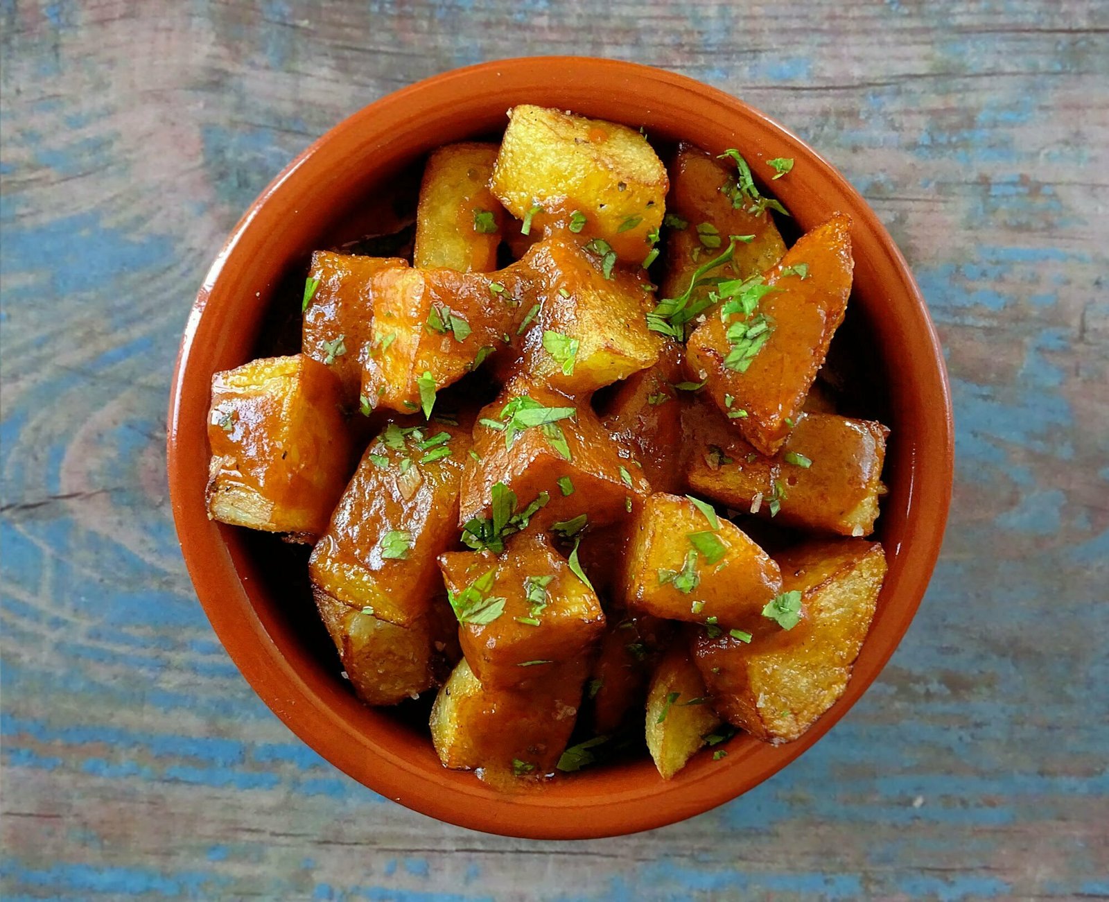 an earthenware dish sits on a blue wooden table and is filled with patatas bravas (golden friend potato pieces smothered with Spanish bravas sauce)