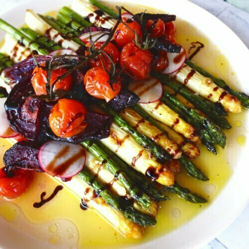 A stack of greenand white asparagus sits on a large white plate with cherry tomatoes, roasted beets and radishes for a garnish