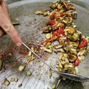 tomato, onion, artichoke, and garlic are stirred on a large paella pan