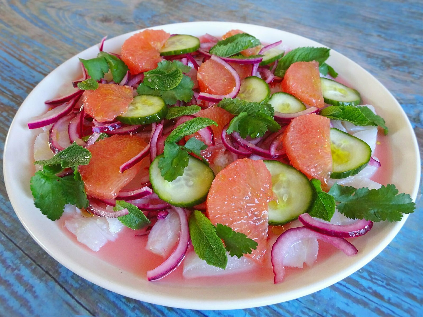 White fish ceviche sits on a white plate garnished with finely sliced red onion, pink grapefruit wedges, and some sprigs of mint and cilantro