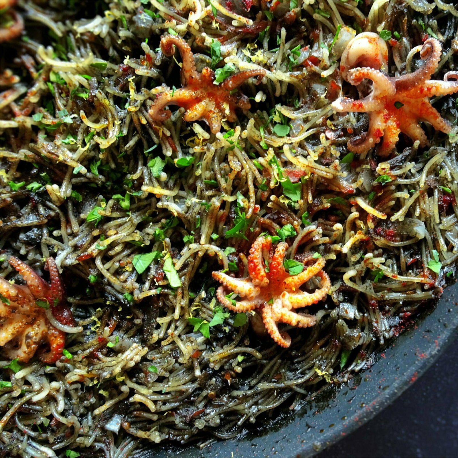 A alrge paella pan is filled with fideua negra and topped with bright red baby squid and some cilantro