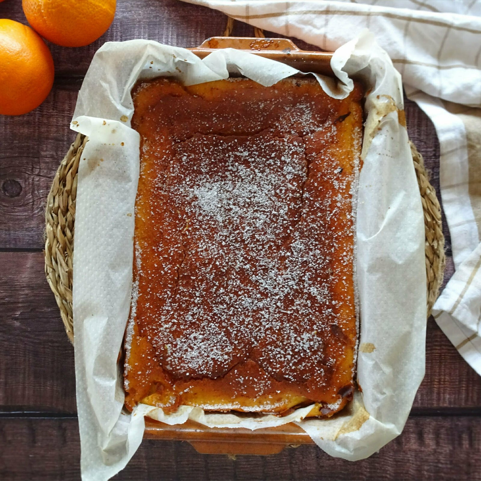 An earthenware dish sits on a rustic wooden counter with a freshly baked orange and pink grapefruit cake covered in baking paper.