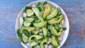 Sliced avocado, cucumber, and radishes sits on a bed of lambs lettuce on a white plate