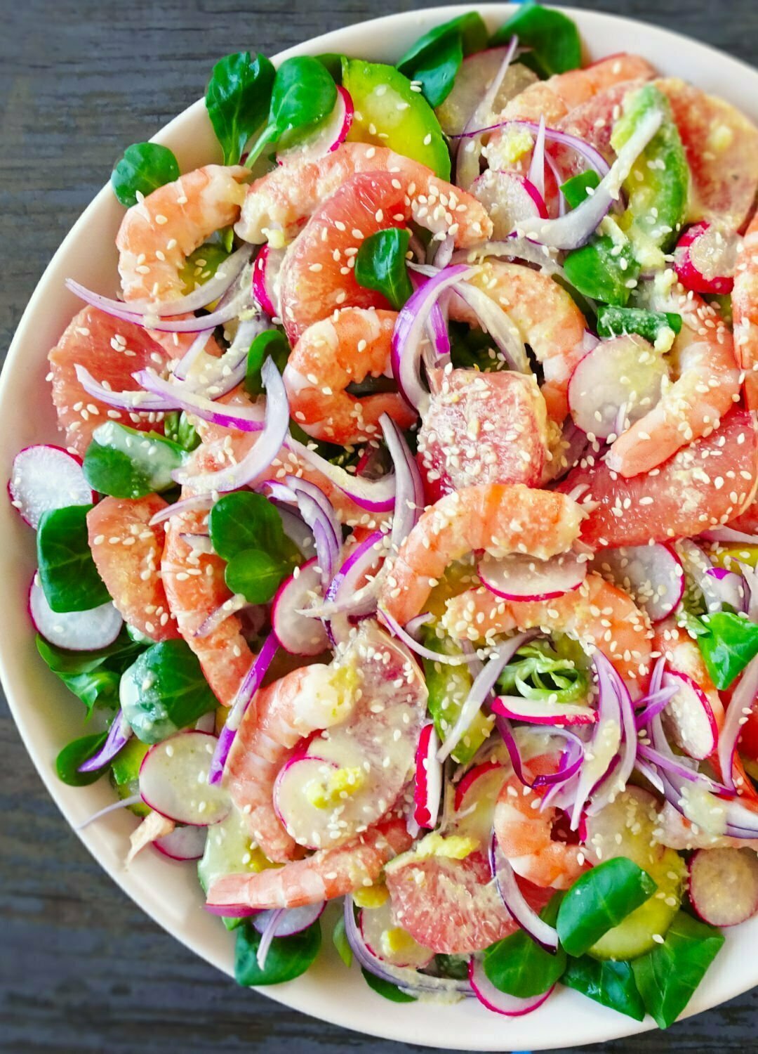 A colorful shrimp and avocado salad sits on a white plate with bright red radishes, pink grapefruit, red onion, and cooked shrimp