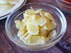 a small glass ramikin sits on a wooden board filled with sliced garlic