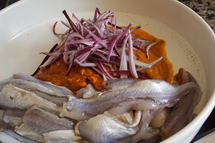 Some fish fillets, sliced red onion, and romesco sauce sit in a casserole dish waiting to be mixed. 