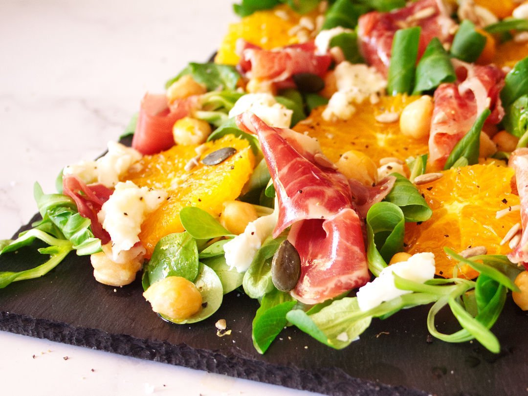 A large platter of jamon iberico sald sits topped with mozarella, salad greens, chickpeas, and seasoned with salt and pepper