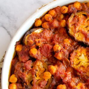 A large white casserole dish sits on a white marble counter with a rich red Mediterranean Eggplant casserole with chickpeas and tomato