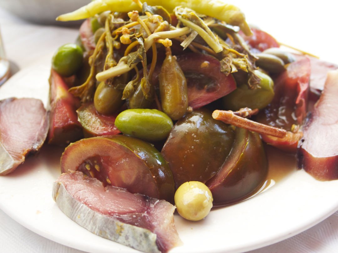 A traditional Murcian salad sits on a plate with some slices of raw tuna, olive, ripe black tomatoes, and giant capers