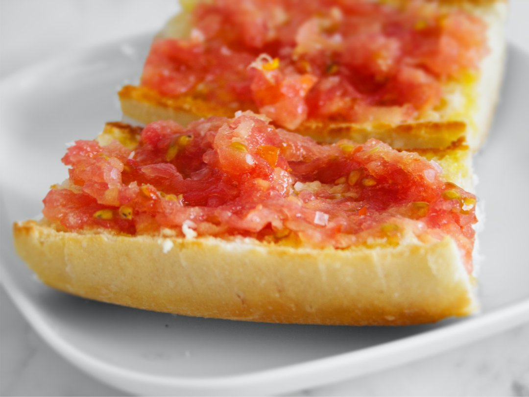 a white plate sits on a marble counter. with two slices of pan con tomate.