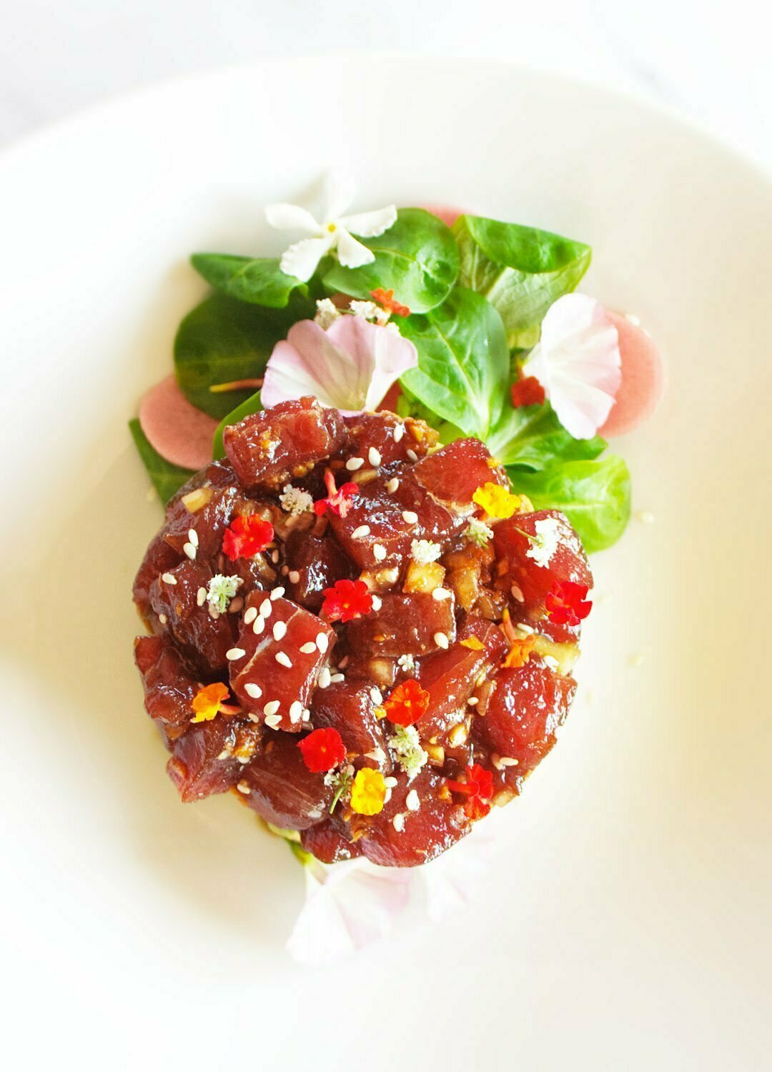 An overhead angle of a stack of tuna tartare is built on top of some avocado and garnished with some green salad leaves and edible flowers