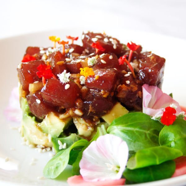 A stack of tuna tartare is built on top of some avocado and garnished with some green salad leaves and edible flowers