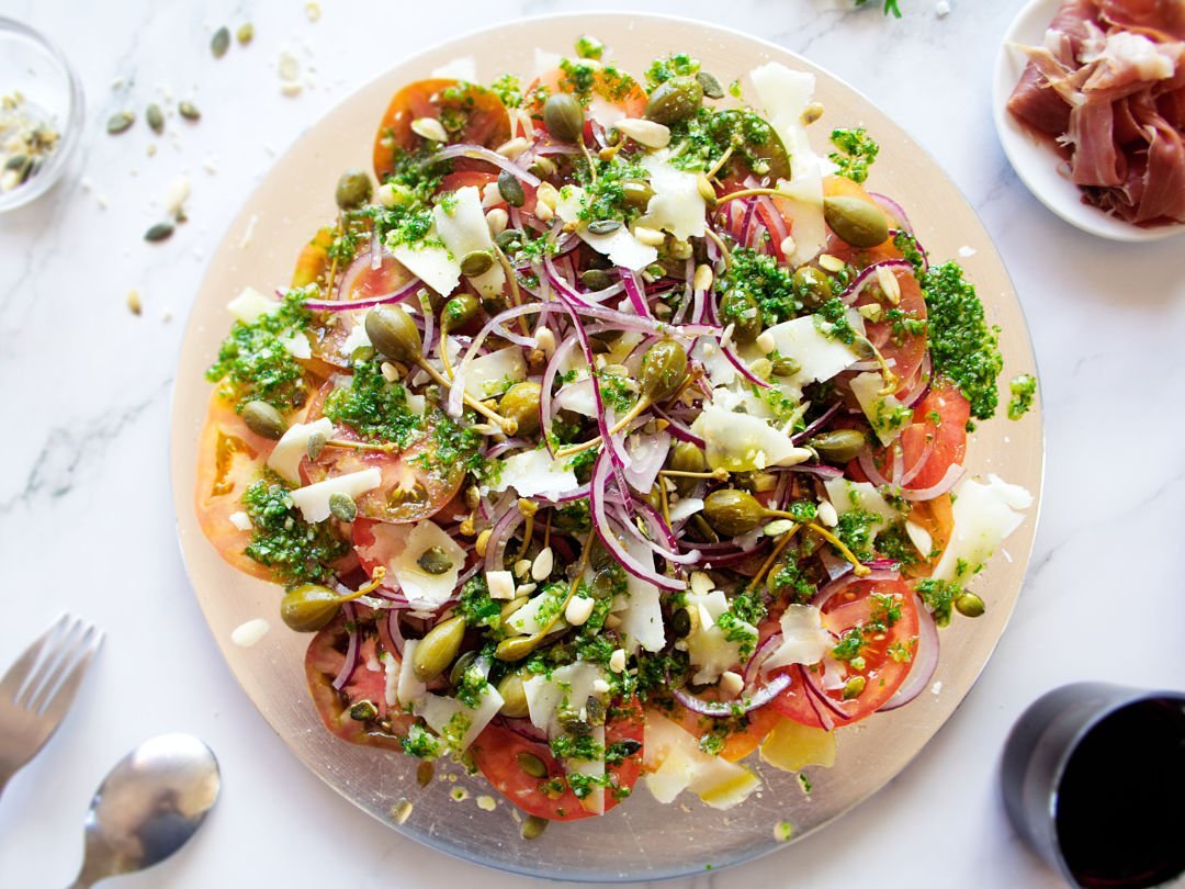 A large plate of Mediterranean Tomato Salad sits. with lots of pesto dressing and shaved pieces of Manchego cheese