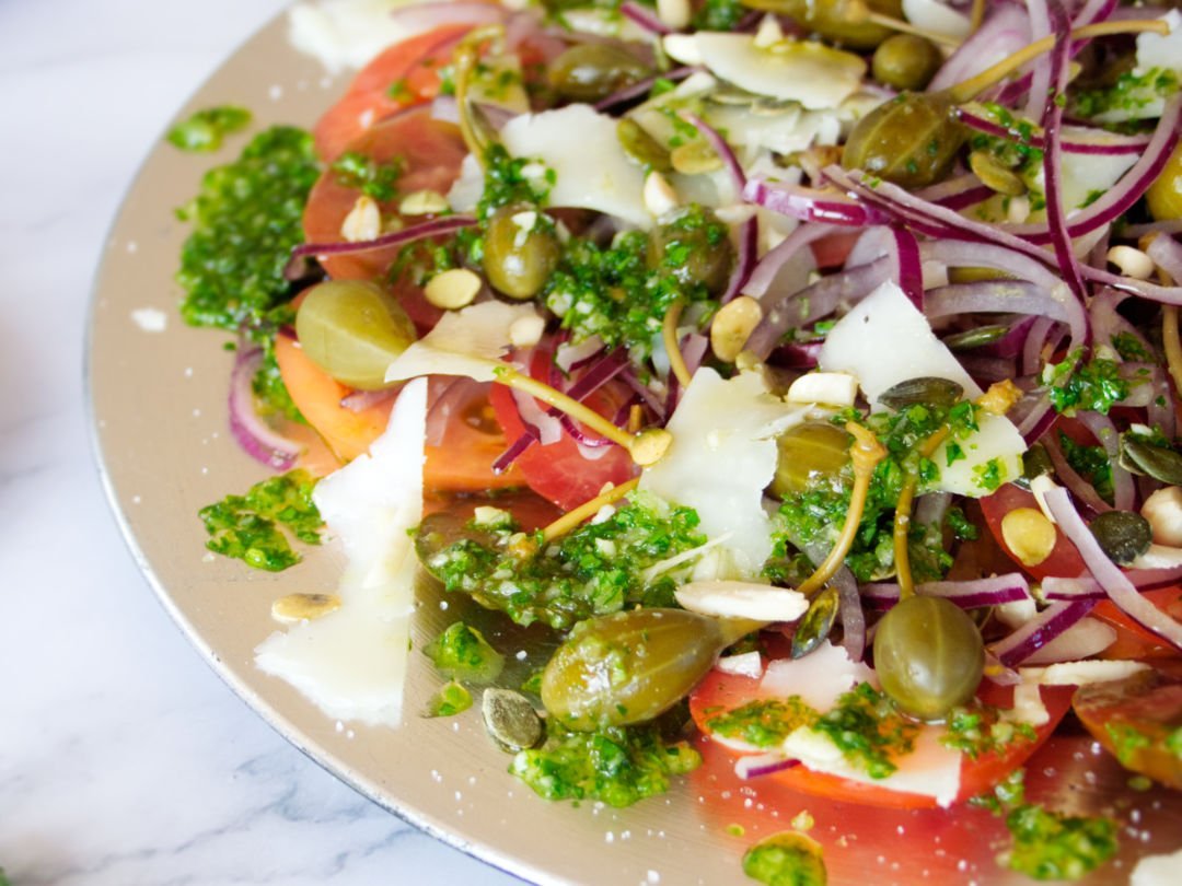 A large plate of Mediterranean Tomato Salad sits. with lots of pesto dressing and shaved pieces of Manchego cheese