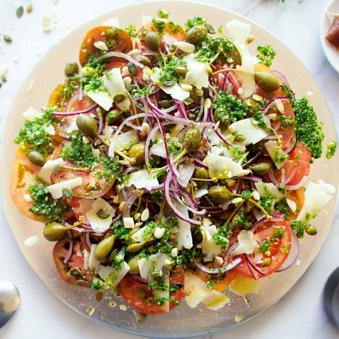 A large plate of Mediterranean Tomato Salad sits. with lots of pesto dressing and shaved pieces of Manchego cheese