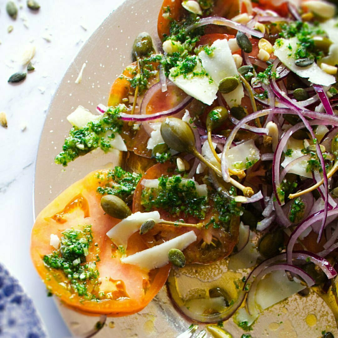 A large platter of Mediterranean Tomato Salad sits drizzled with homemade pesto dressing and some flakes of Machego cheese 