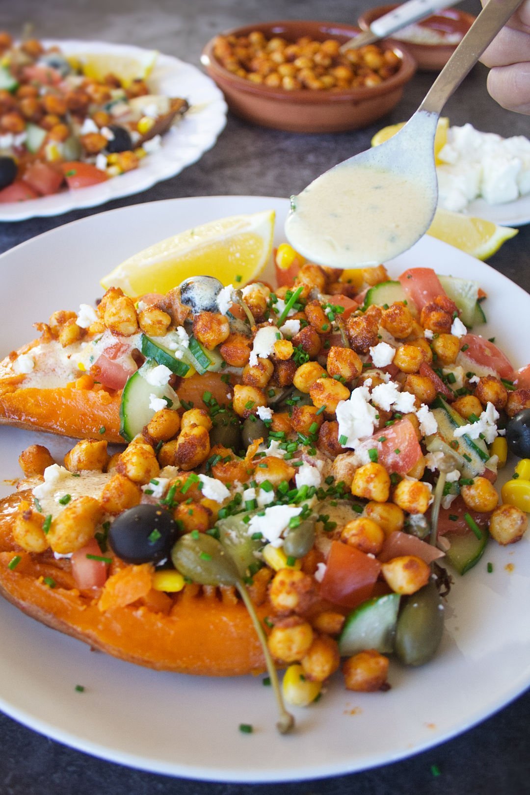 a large baked sweet potato sits on a white plate with plenty of Mediterranean inspired toppings.