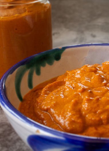 A small bowl of romesco sauce sits with a jar full of romesco sauce in the background