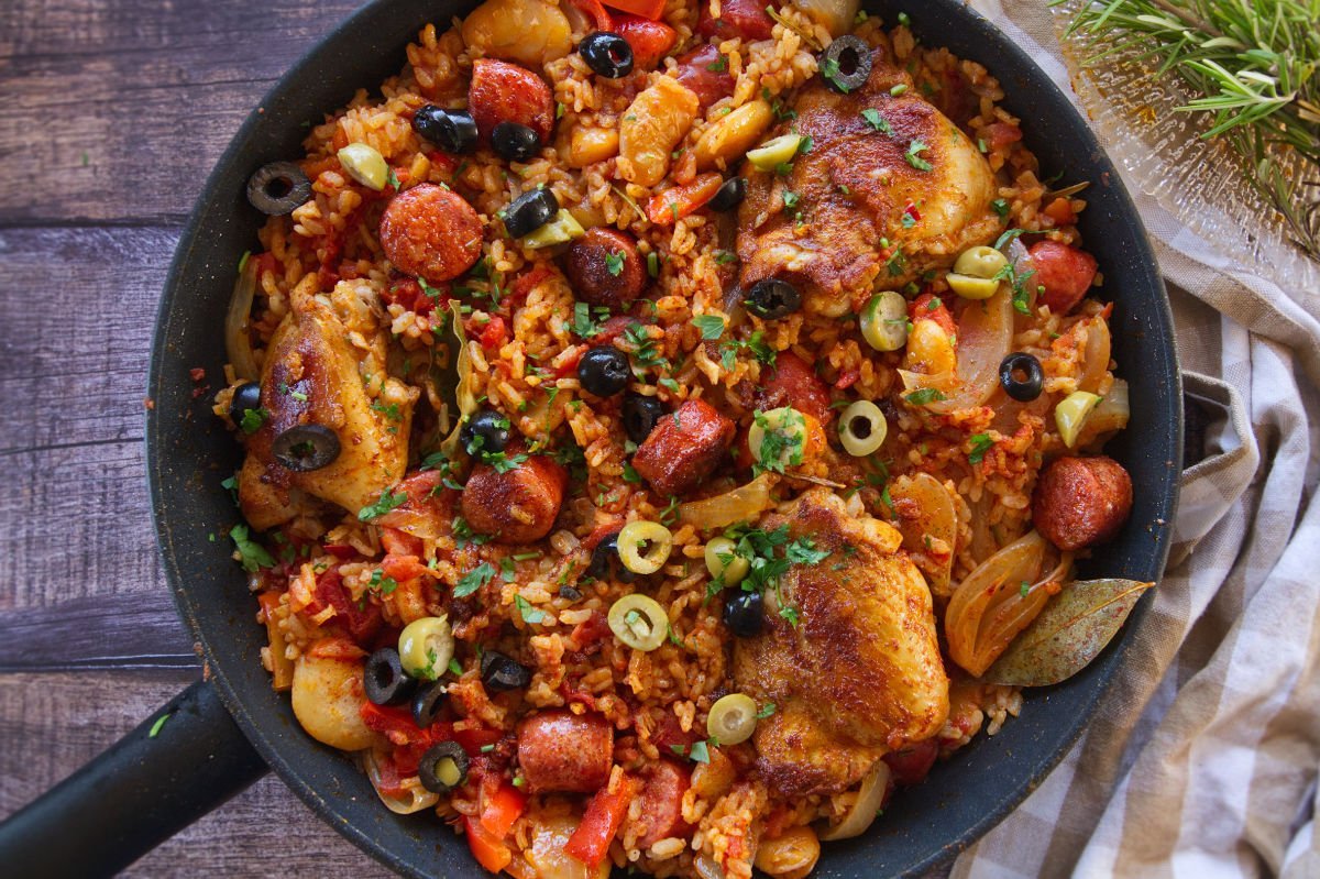 A large pan of Spanish chicken and rice sits on a counter beside some fresh rosemary