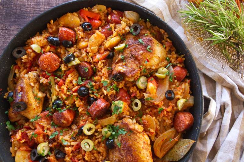 A large pan of Spanish chicken and rice sits garnished with olives and fresh chopped parsley