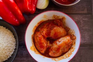 A few pieces of marinated chicken sit in a bowl beside some fresh peppers and rice