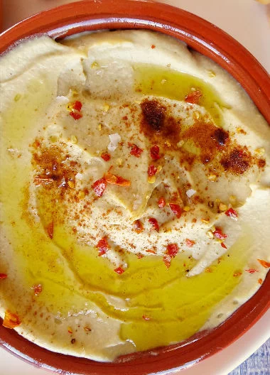 A small bowl of Mediterranean hummus is garnished with some olive oil, salt, paprika, and chili flakes