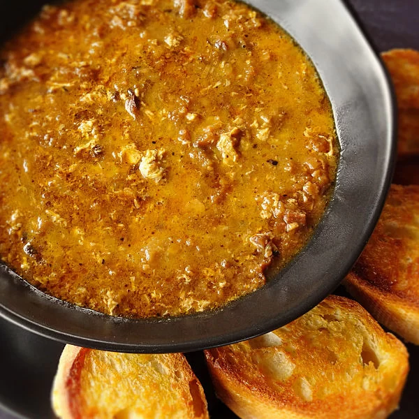 A steaming bowl of garlic soup sits in a black bowl beside a few slices of golden toasted bread