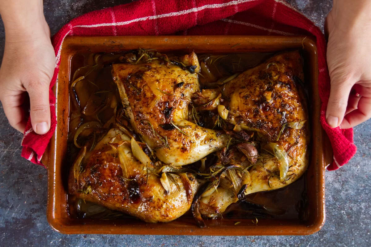 a person serves a large tray. of pollo al horno beside somw fresh sprigs of rosemary