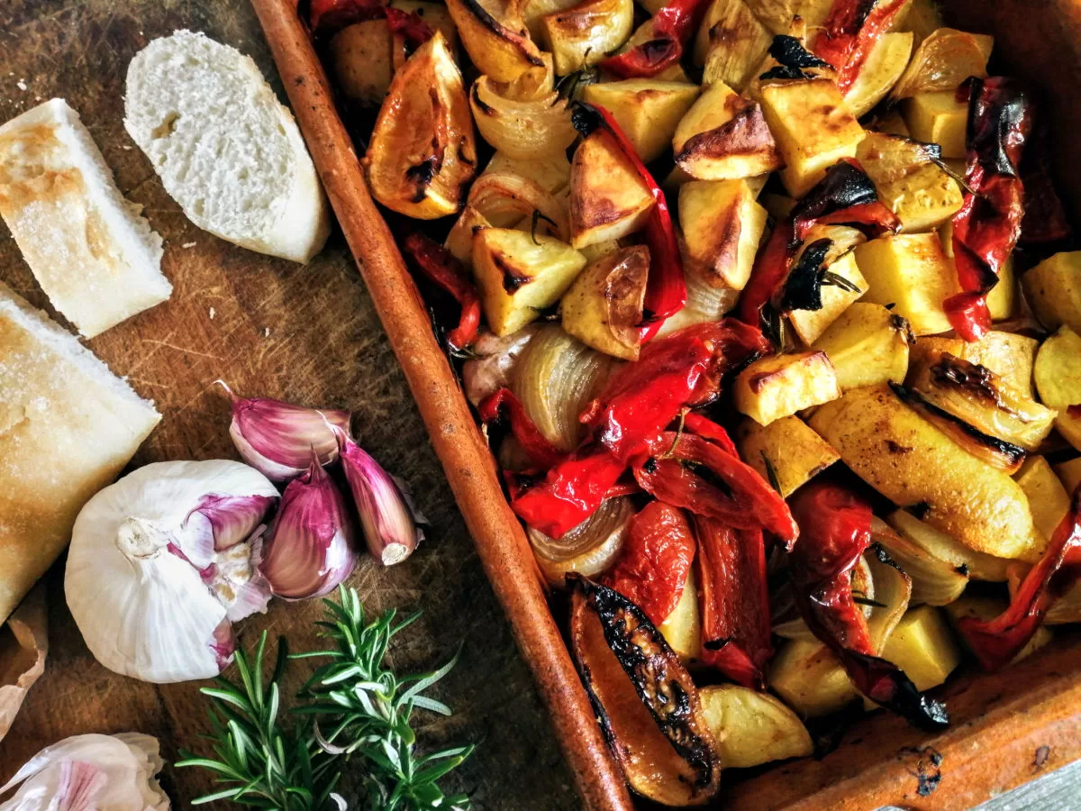 A large tray of poor man's potatoes sits beside some fresh cloves of garlic and herbs