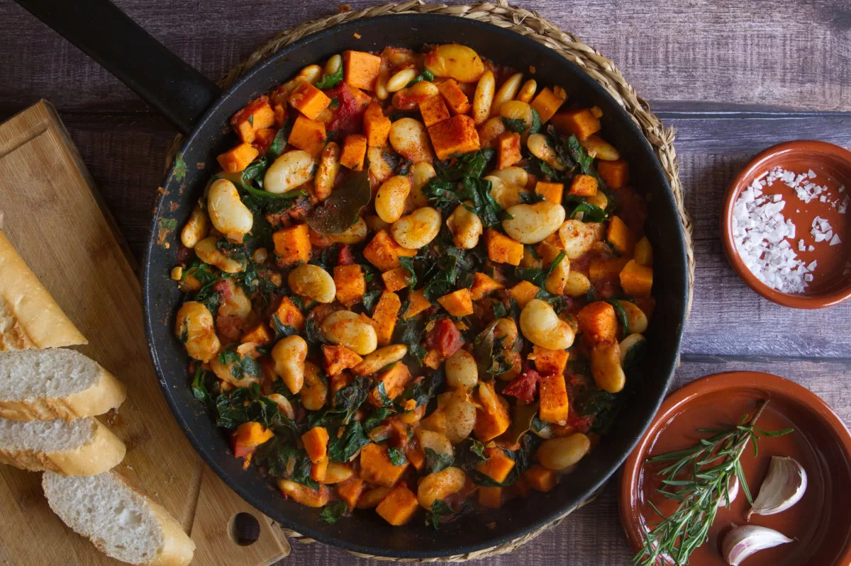 A large pan of smoky butter bean stew sits beside some sliced bread and crushed seas salt. 