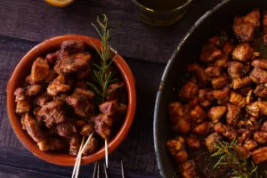 a small bowl of paprika-infused pork bites sits next to. alrge fry pan of cooked pork.