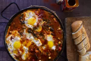 a pan of chickpeas with spinach and eggs sits beside some sliced bread.