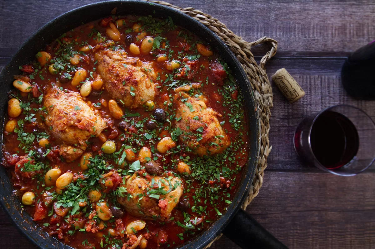 A pan of Spanish chicken thighs with chorizo and wine sits beside a small glass of red wine.