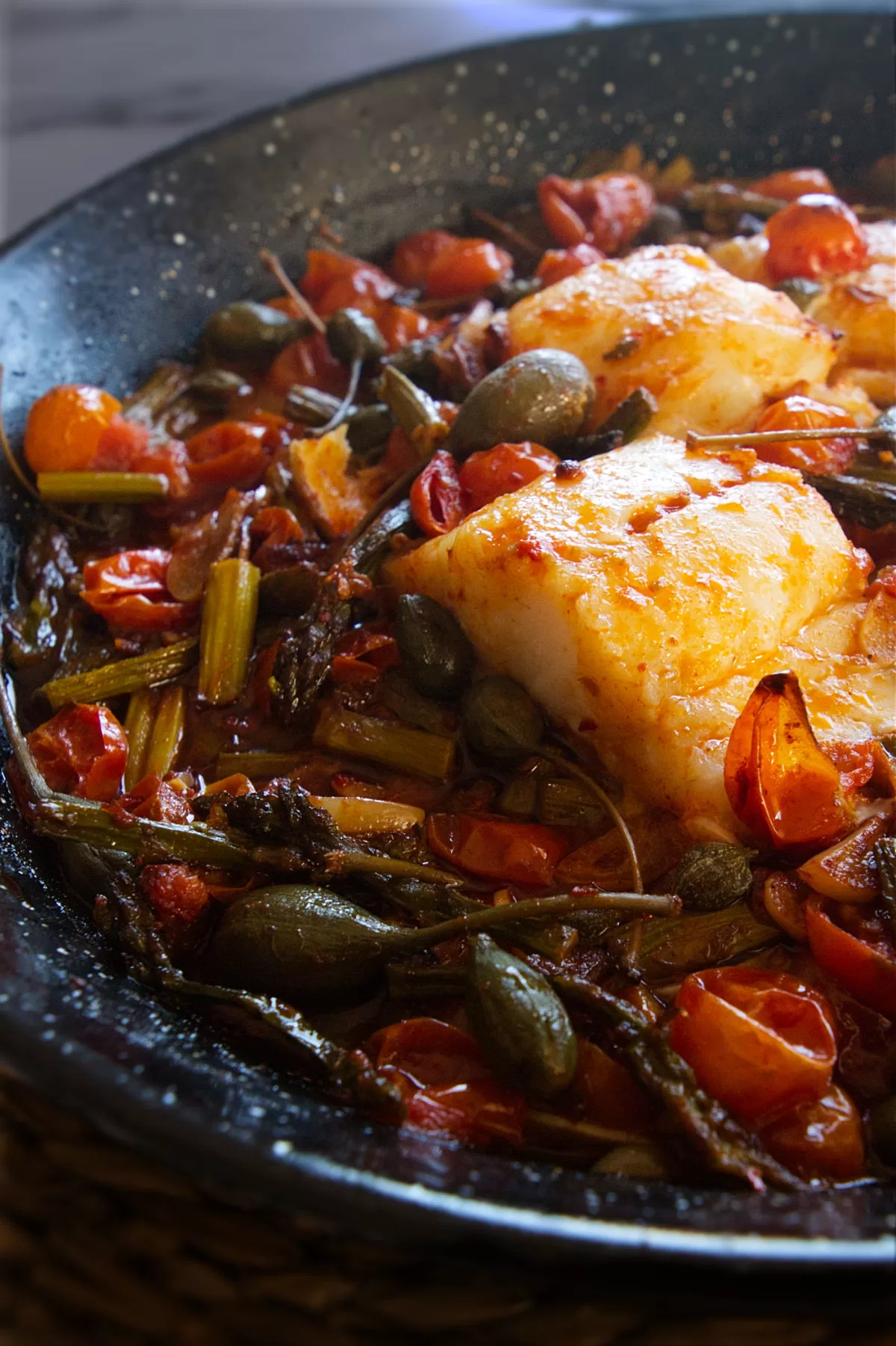 a few fillets of cod fish sit in a pan with sauteed asparagus and cherry tomatoes.