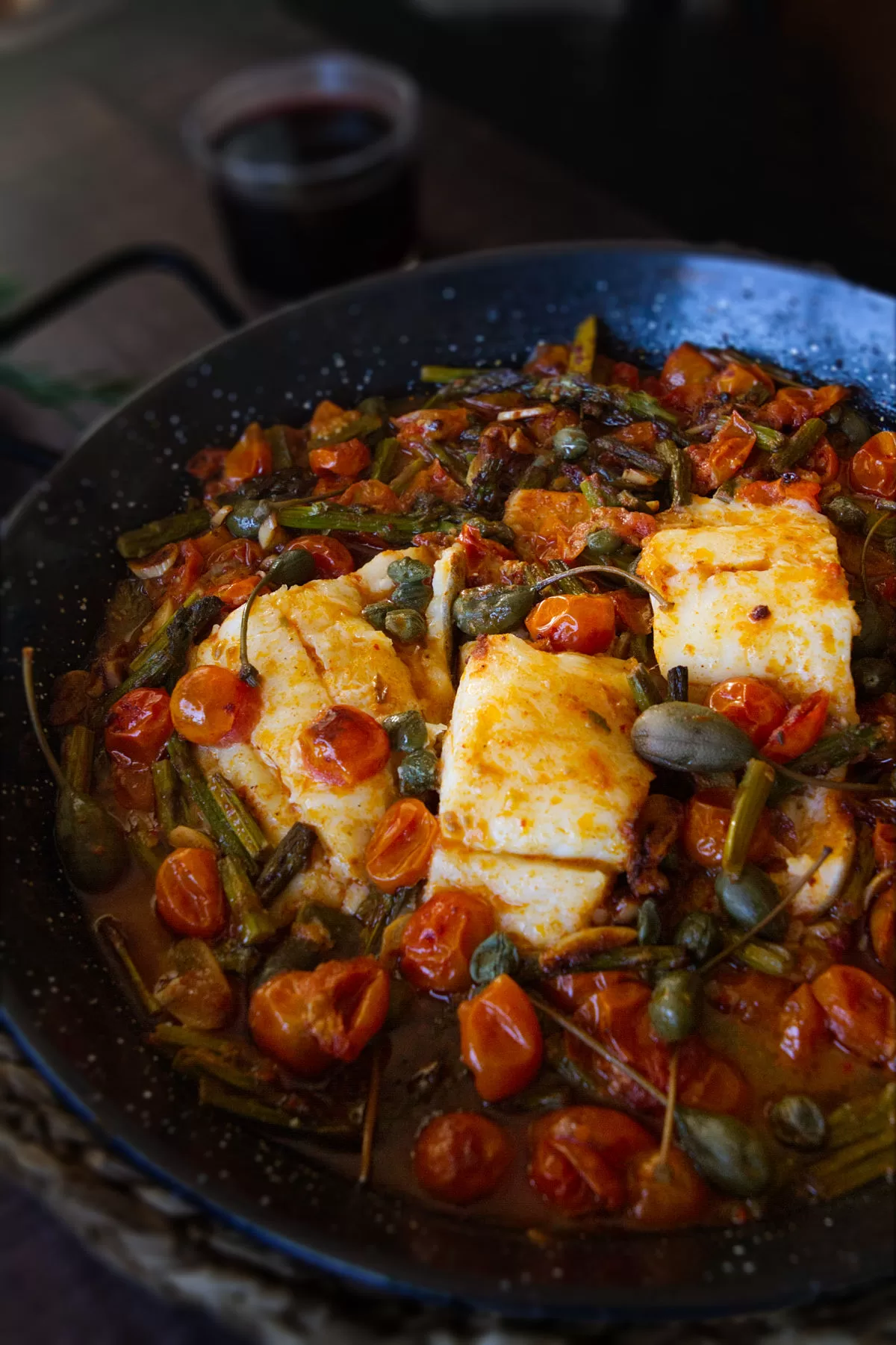 a few fillets of cod fish sit in a pan with sauteed asparagus and cherry tomatoes.