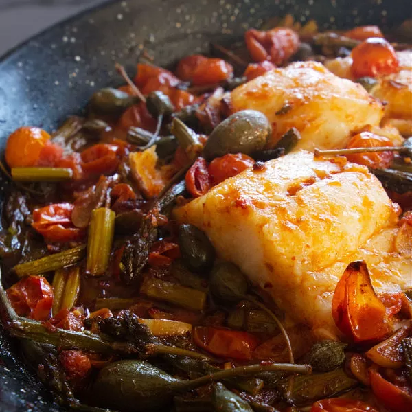 a few fillets of cod fish sit in a pan with sauteed asparagus and cherry tomatoes.