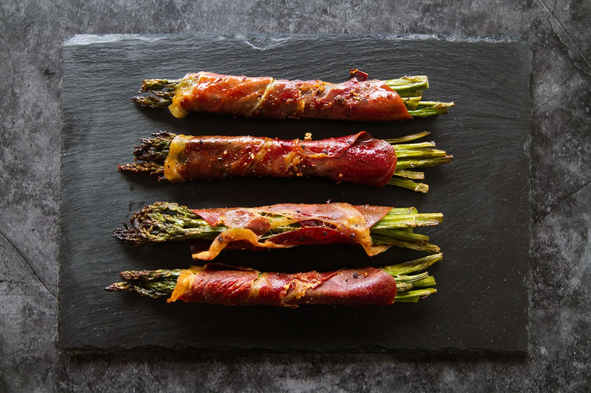 Roasted asparagus spears wrapped in Serrano ham sit on a slate plate.