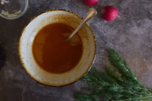 a bowl of salad dressing sits beside some fresh dill and radishes.