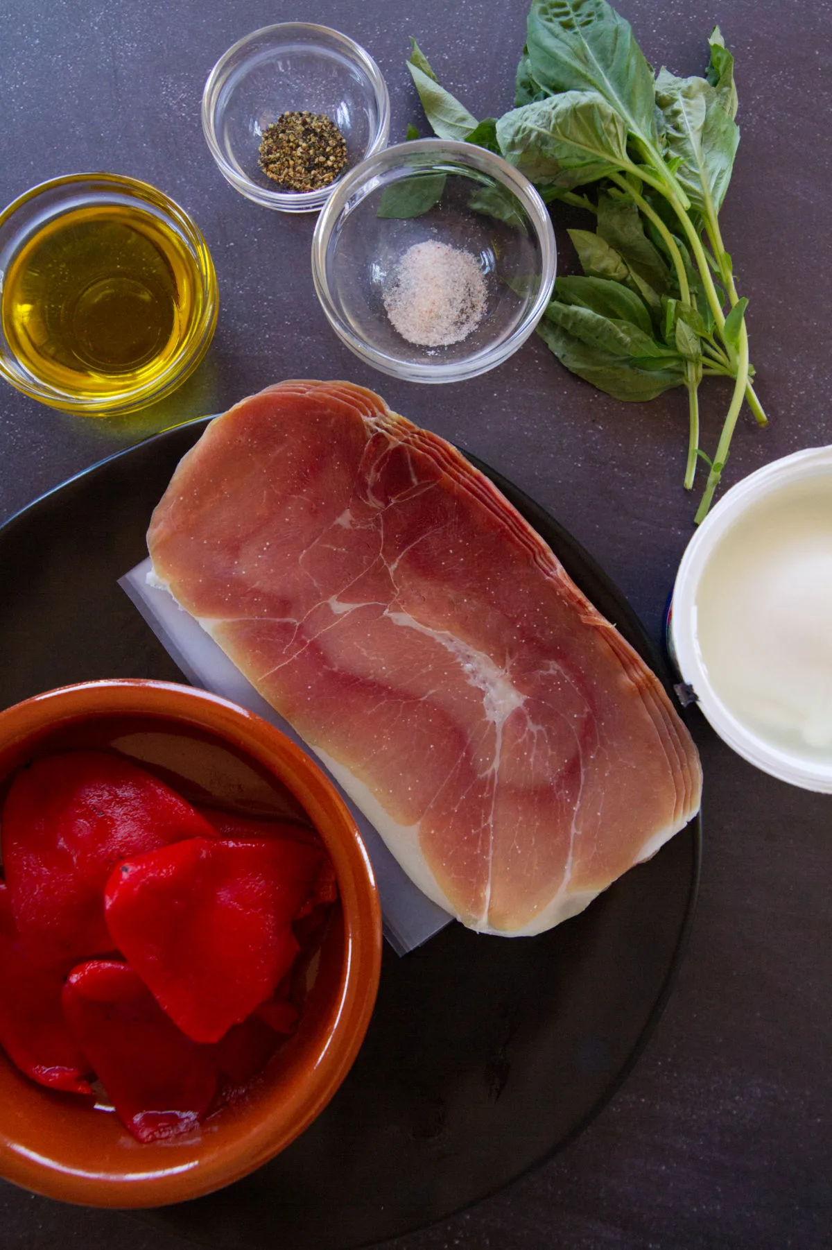 Burrata with Serrano ham and roast pepper ingredients sit beside fresh basil and some salt and pepper. 