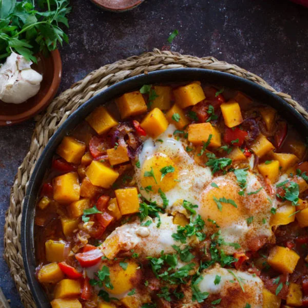 A large pan of pumpkin pisto sits beside some brown bread and fresh parsley.